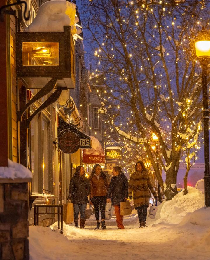 Four people walking down a snow-covered sidewalk at night, store window and streetlights lit up 