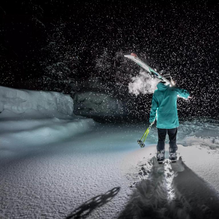 A skier holding their ski's over their right shoulder and ski poles in their left hand at night and snow falling from the sky