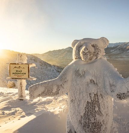 A statue of a bear covered in snow at the top of a mountain with a sign that reads Bear Mountaiin behind it