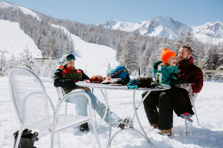 Two adults enjoying a beer in front of the slopes while a child is laughing. 