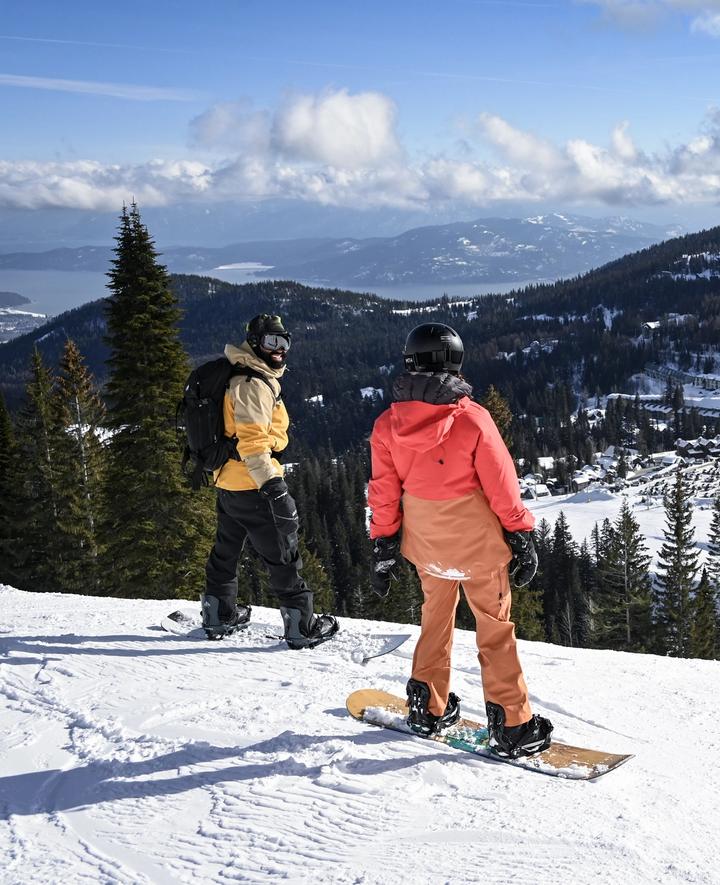 Two people on snowboards standing at the top of a run looking down at a mountain village