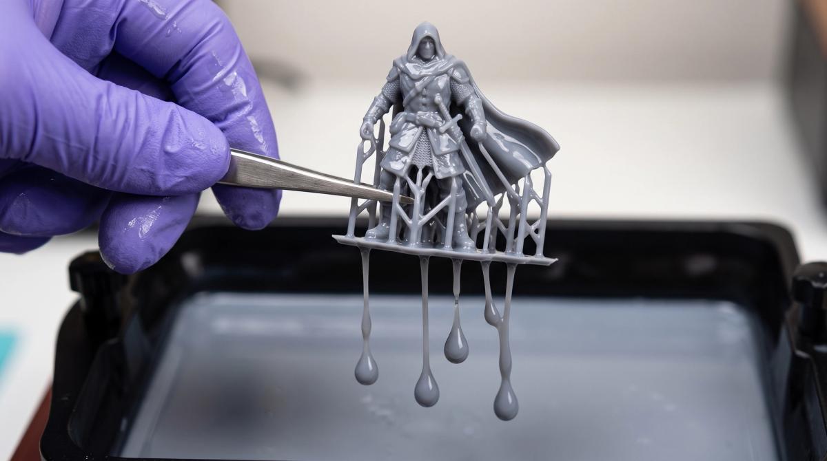 Hands wearing purple nitrile gloves holding a grey resin miniature dripping with excess uncured resin over an open vat