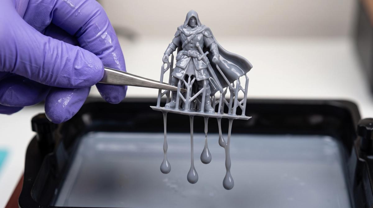 Hands wearing purple nitrile gloves holding a grey resin miniature dripping with excess uncured resin over an open vat