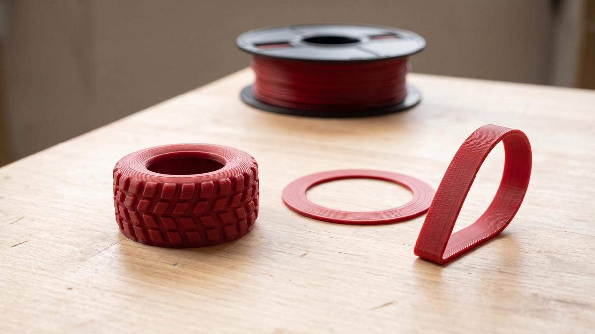 Flexible red TPU prints - a small treaded tire, a flat gasket ring and a strap bent into a loop - next to a spool of the same filament
