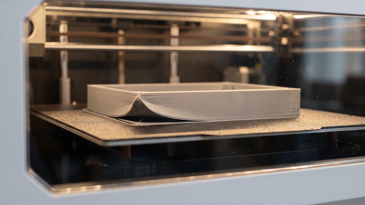 A grey ABS print inside an enclosed printer chamber with one corner visibly warped off the build plate