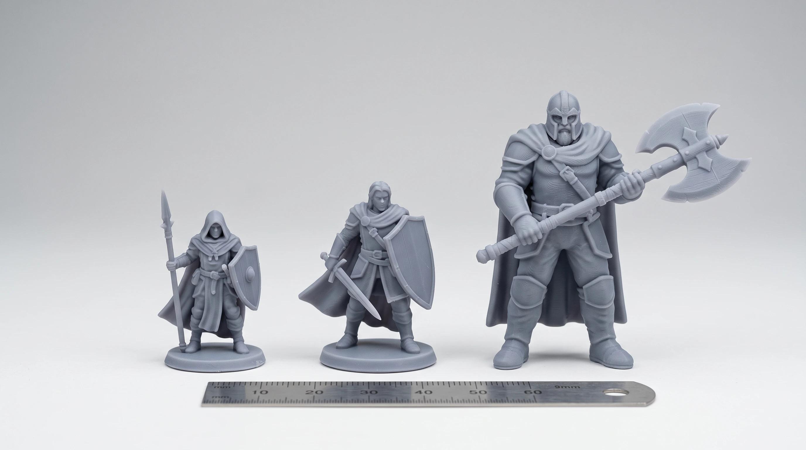 Three grey miniature figures of clearly different heights standing in a row with a measuring ruler in front