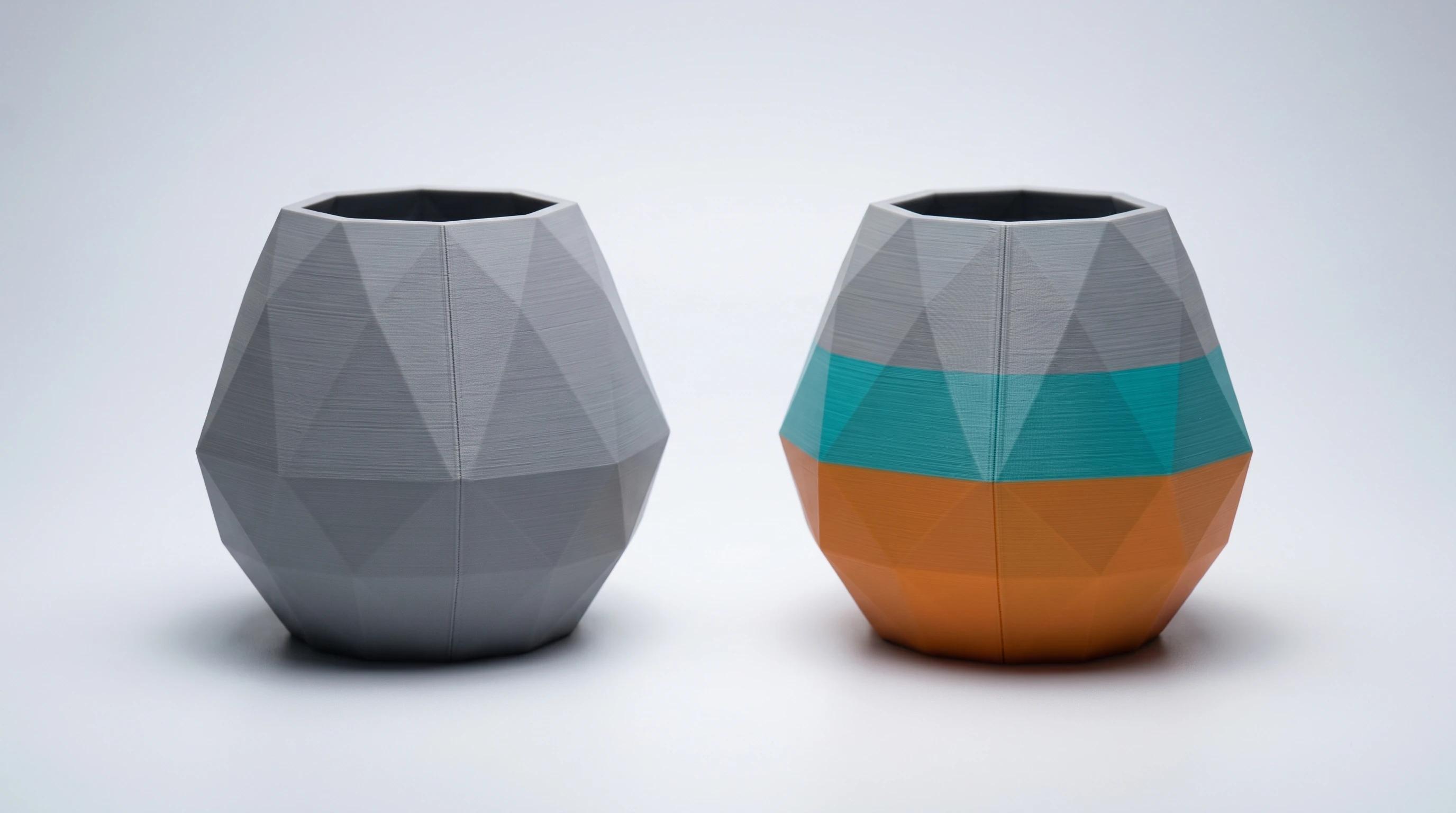 Two identical faceted 3D printed vases side by side, one plain grey and one printed in orange, teal and grey colour bands