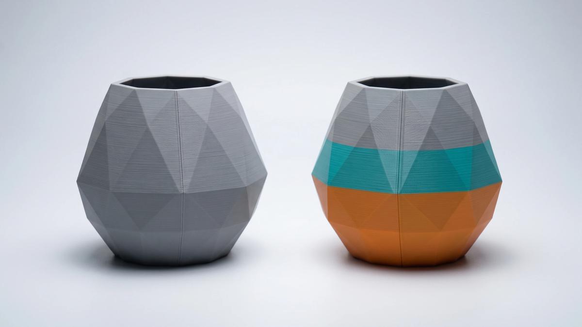 Two identical faceted 3D printed vases side by side, one plain grey and one printed in orange, teal and grey colour bands