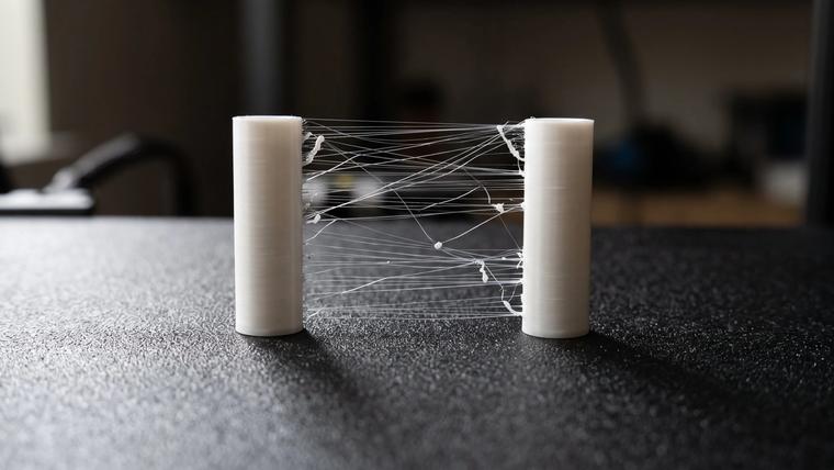 Two white 3D printed towers on a black textured build plate with fine plastic strings and small blobs stretched between them, a classic stringing test