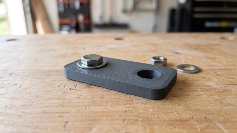 A dark gray 3D printed bracket on a wooden workbench with a steel hex bolt and washer seated in one of its two holes, and a nut and washer lying beside it