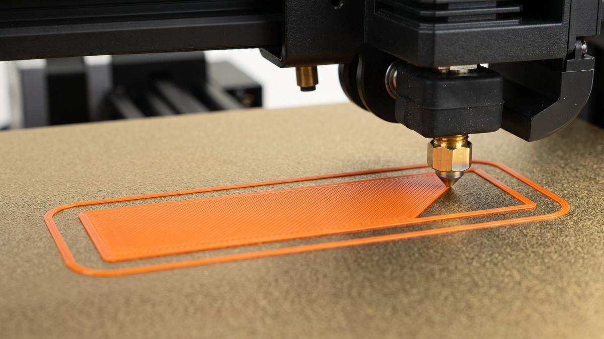 Close-up of a 3D printer nozzle laying the first layer of orange filament on a textured PEI build plate