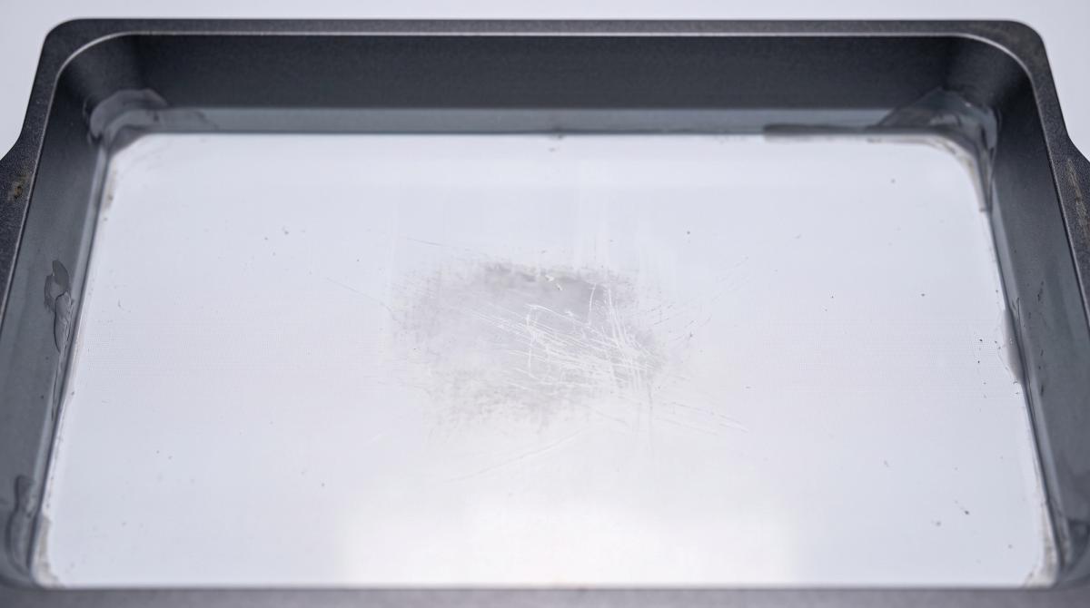 A drained resin vat's film floor showing a scratched, hazy patch of damage next to the clear undamaged film around it