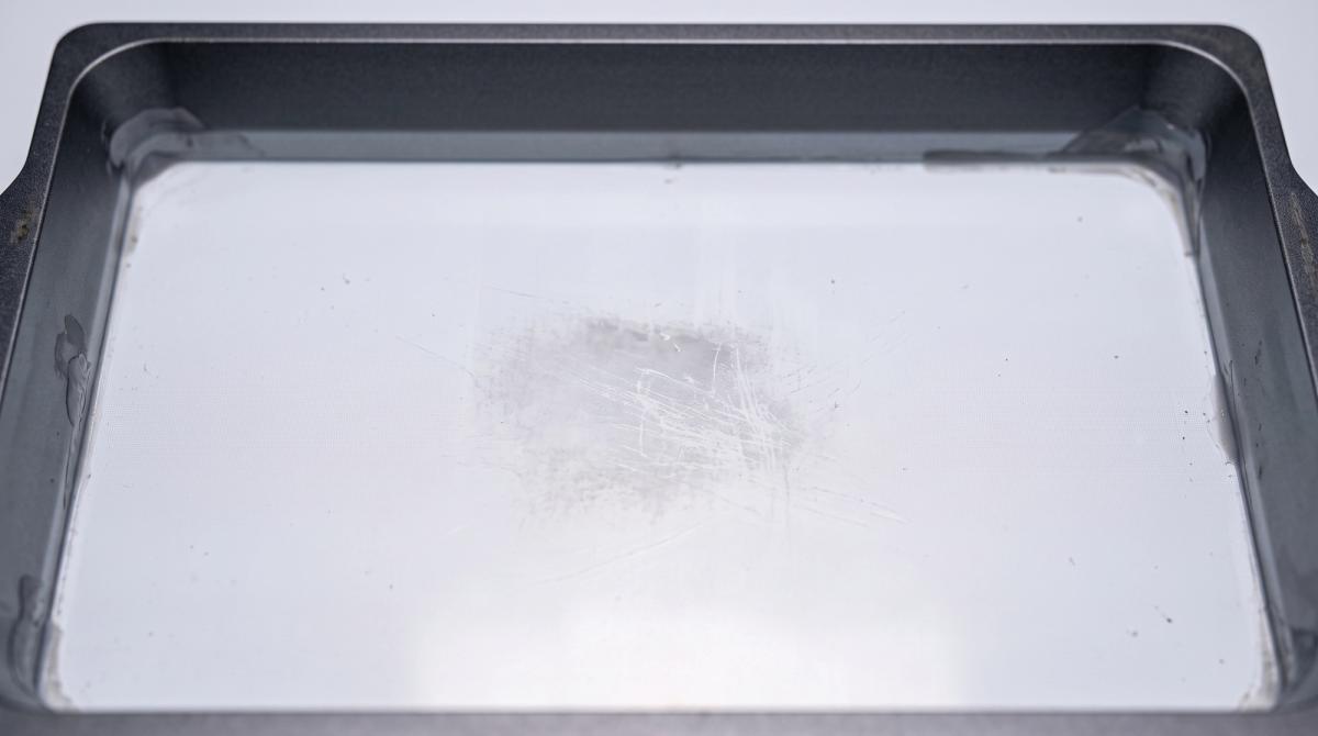 A drained resin vat's film floor showing a scratched, hazy patch of damage next to the clear undamaged film around it