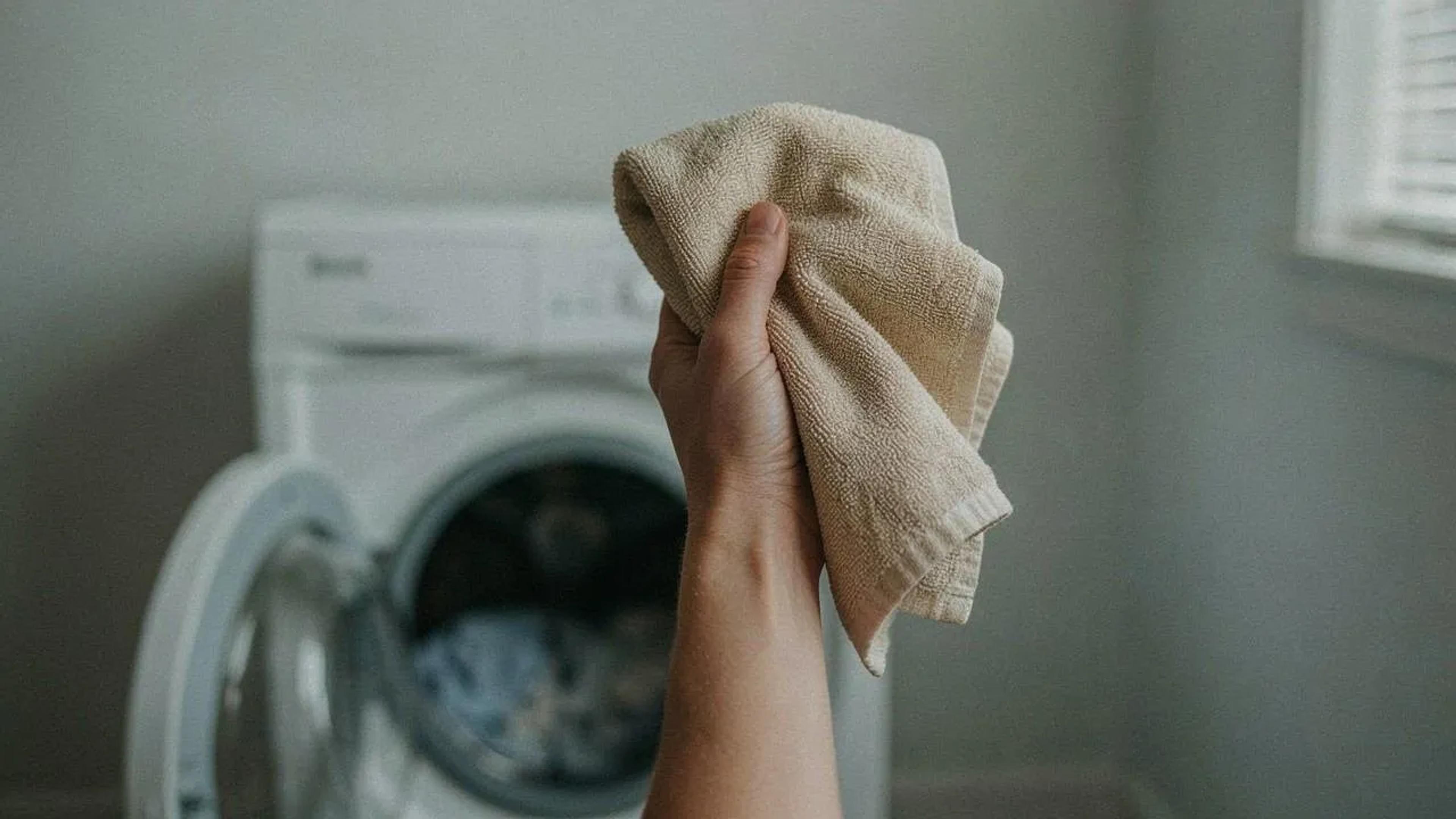 Hand holding up a beige towel in front of an open front-load laundry machine