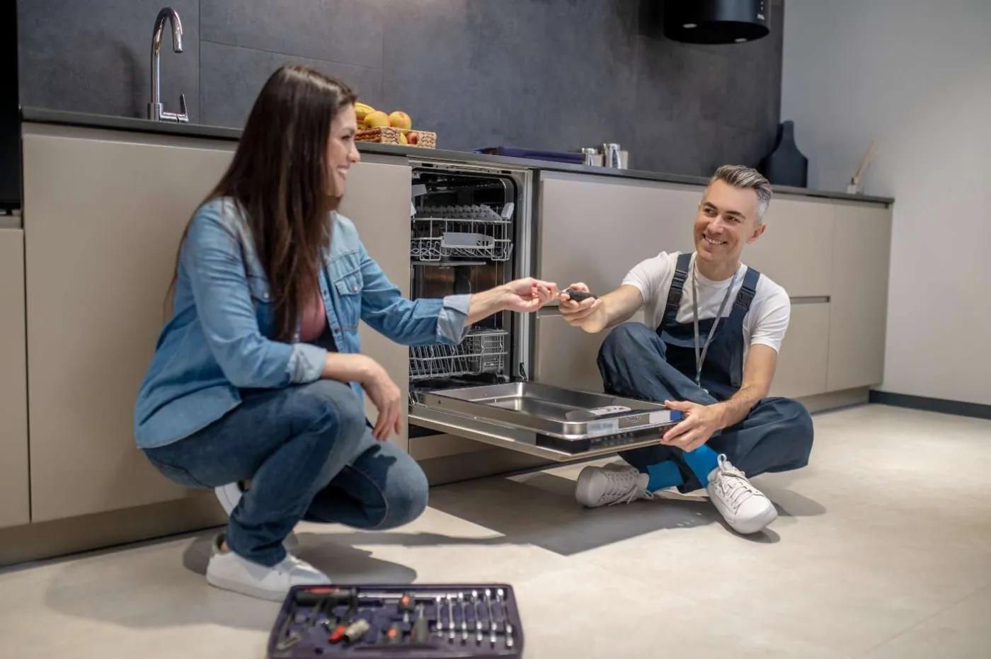 Technician in overalls seated by an open built-in dishwasher, handing a homeowner a screwdriver