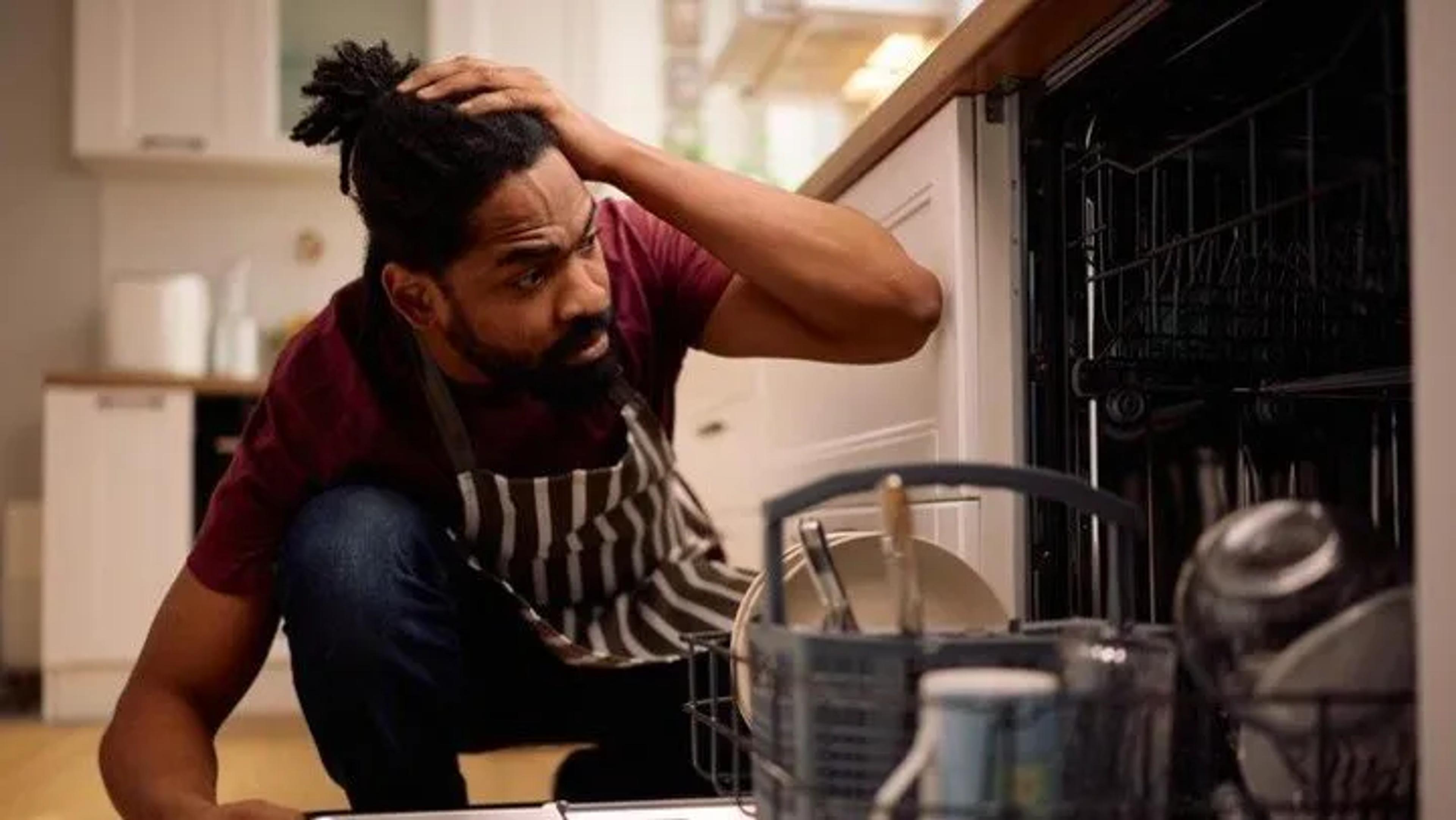 Puzzled man in an apron crouching by an open dishwasher, hand on his head
