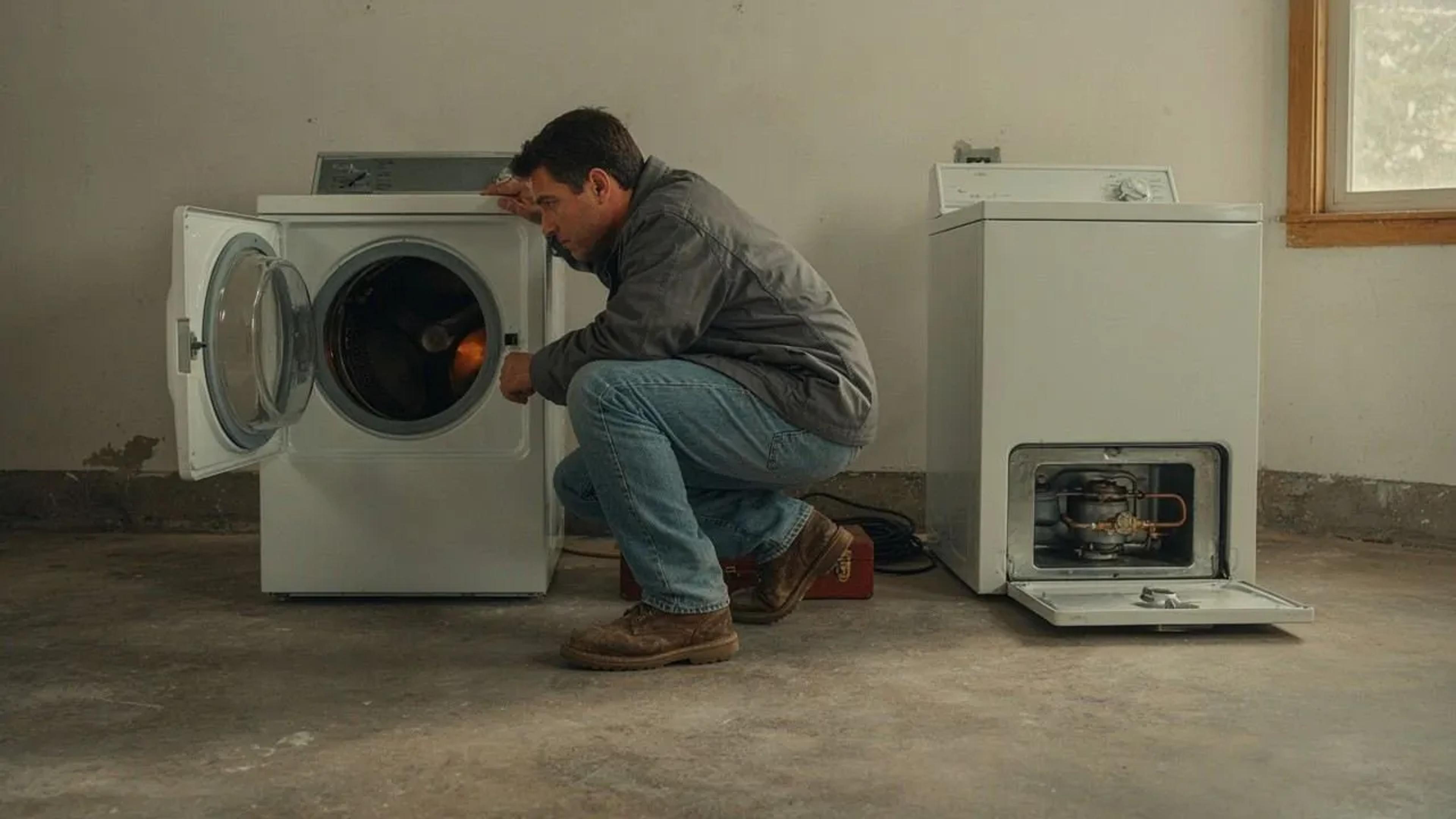 Technician crouching beside an open front-load dryer, with a second dryer showing its gas burner assembly