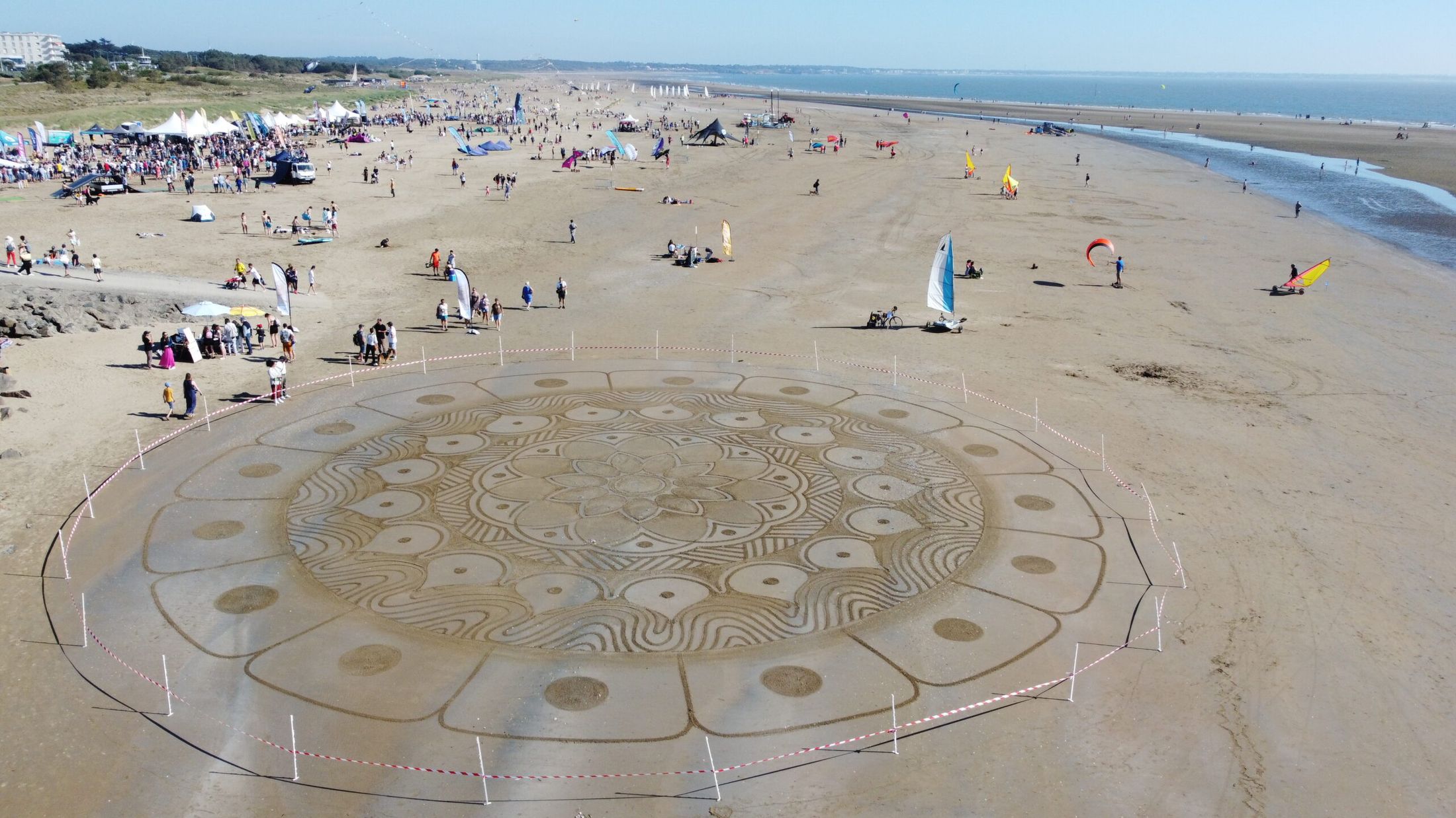 Vue aérienne du Week-end Glisse Saint-Brévin : mandala de sable au premier plan, village des exposants, kites et chars à voile sur la plage de l'Océan. Crédit : Saint-Brévin Tourisme.
