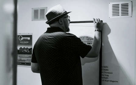 Scott Bennett wearing a fedora and writing on a wall-mounted board at the NOSY studio.