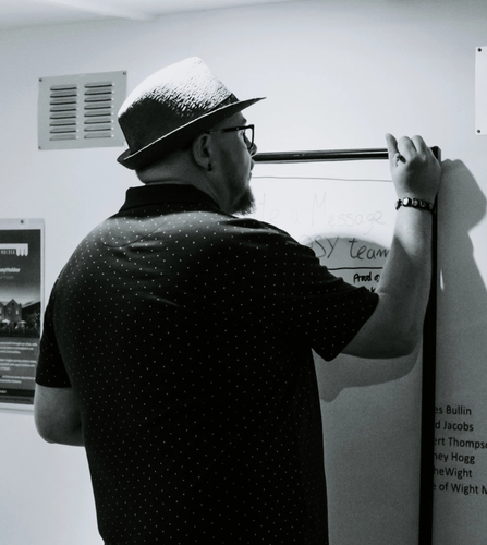 Scott Bennett wearing a fedora and writing on a wall-mounted board at the NOSY studio.