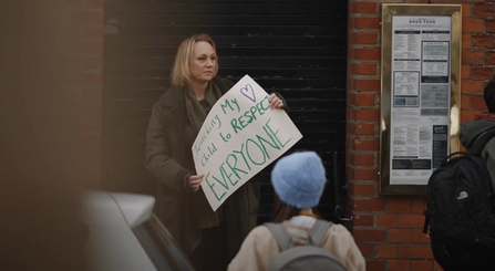 A still of a woman holding a placard reading "Teaching my child to respect everyone"
