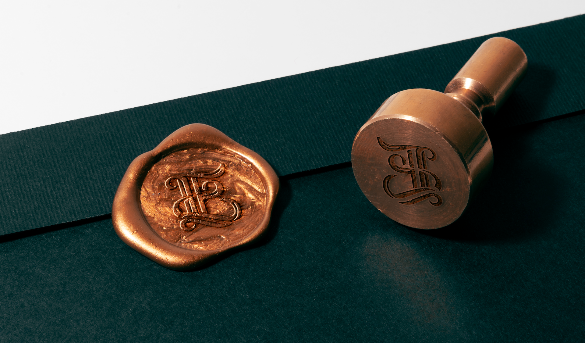 Copper wax seal with monogram logo on a dark envelope beside the engraved stamp tool.