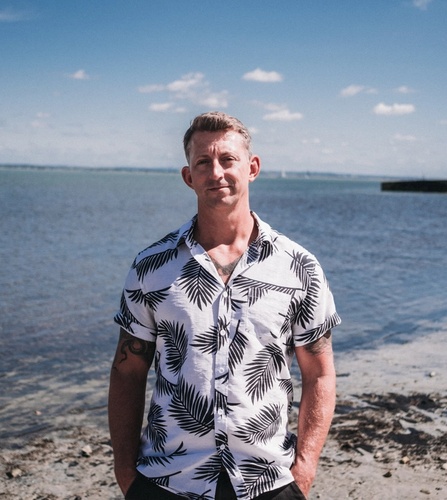 Professional headshot of Dan Carley, Client Director at NOSY Agency, wearing a white shirt with a seaside background.