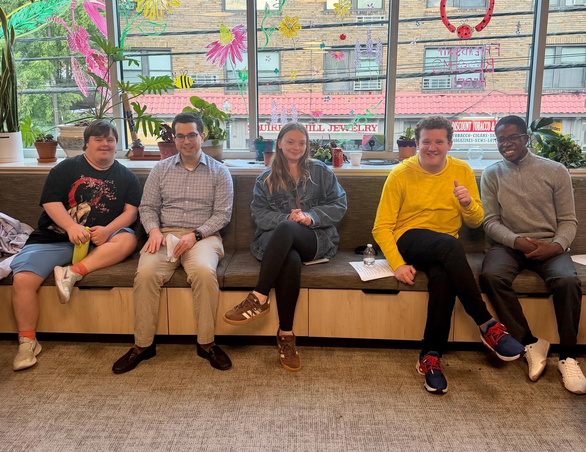 Five Friendship Circle speakers sit together in a bright room with colorful window drawings behind them.