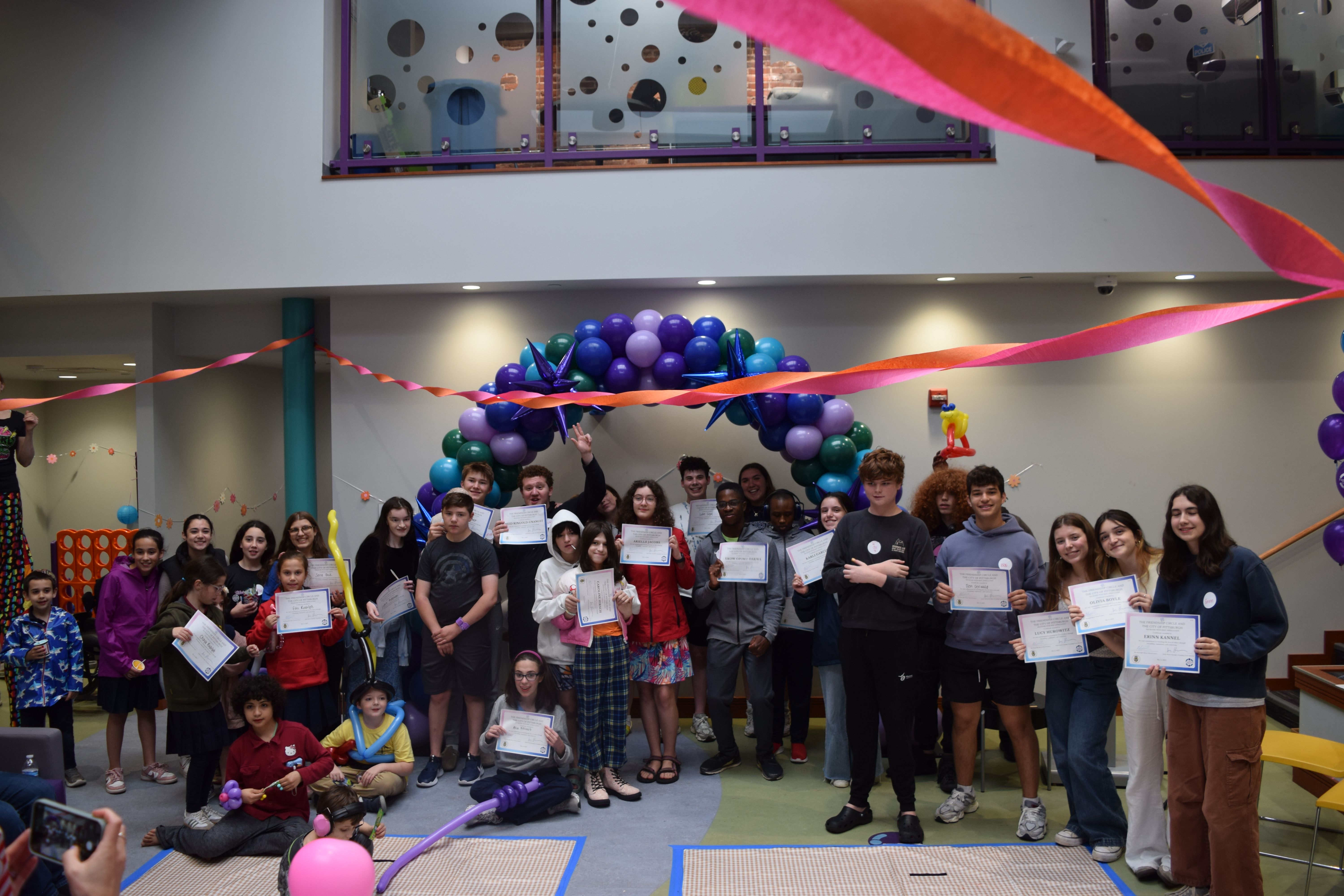 A group photo of boys and girls of all ages smiling, sitting, and standing in front of a blue and purple ballon arch. They are holding up certificates at the Friendship Awards program.