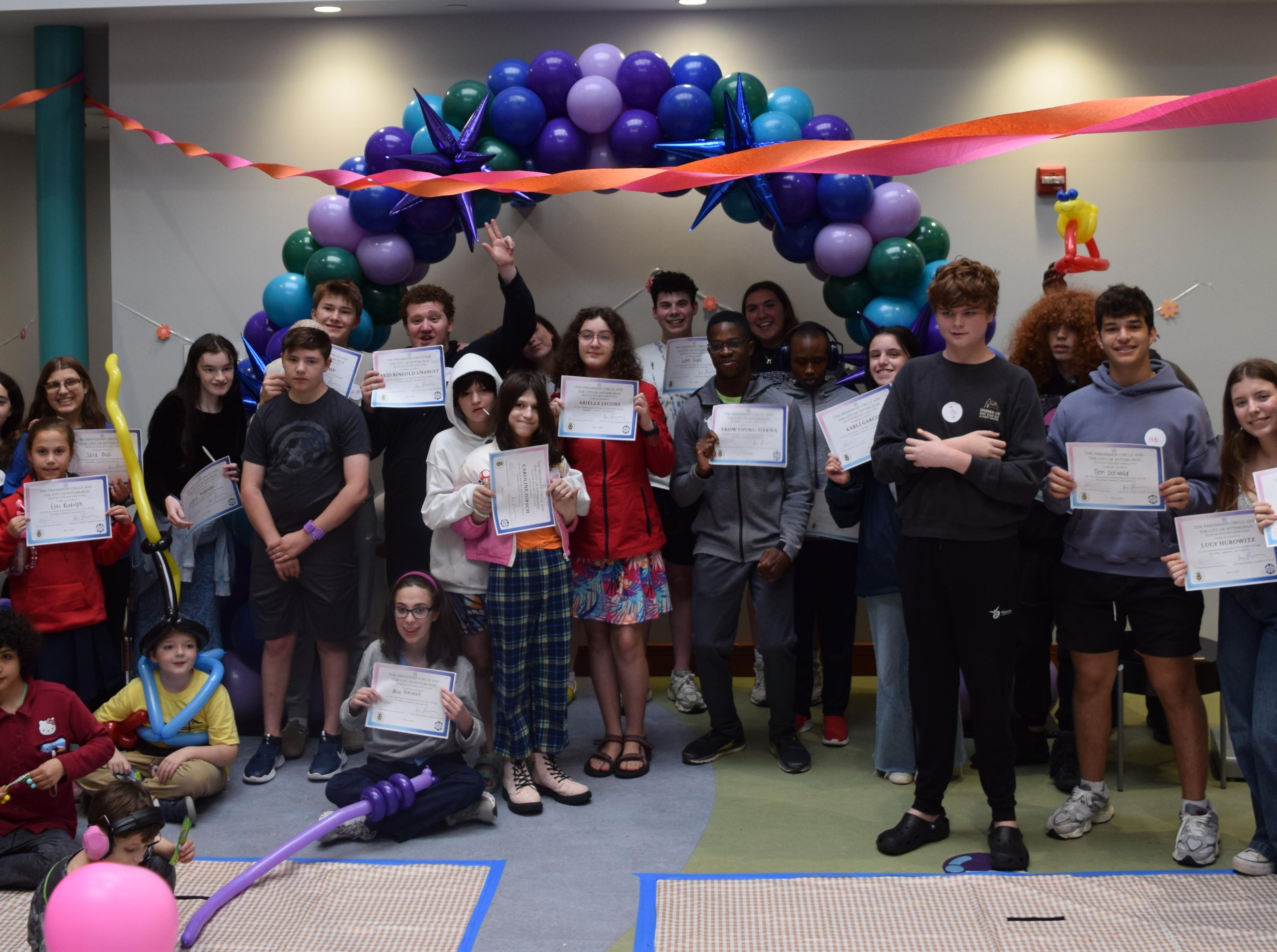 A group photo of boys and girls of all ages smiling, sitting, and standing in front of a blue and purple ballon arch. They are holding up certificates at the Friendship Awards program.