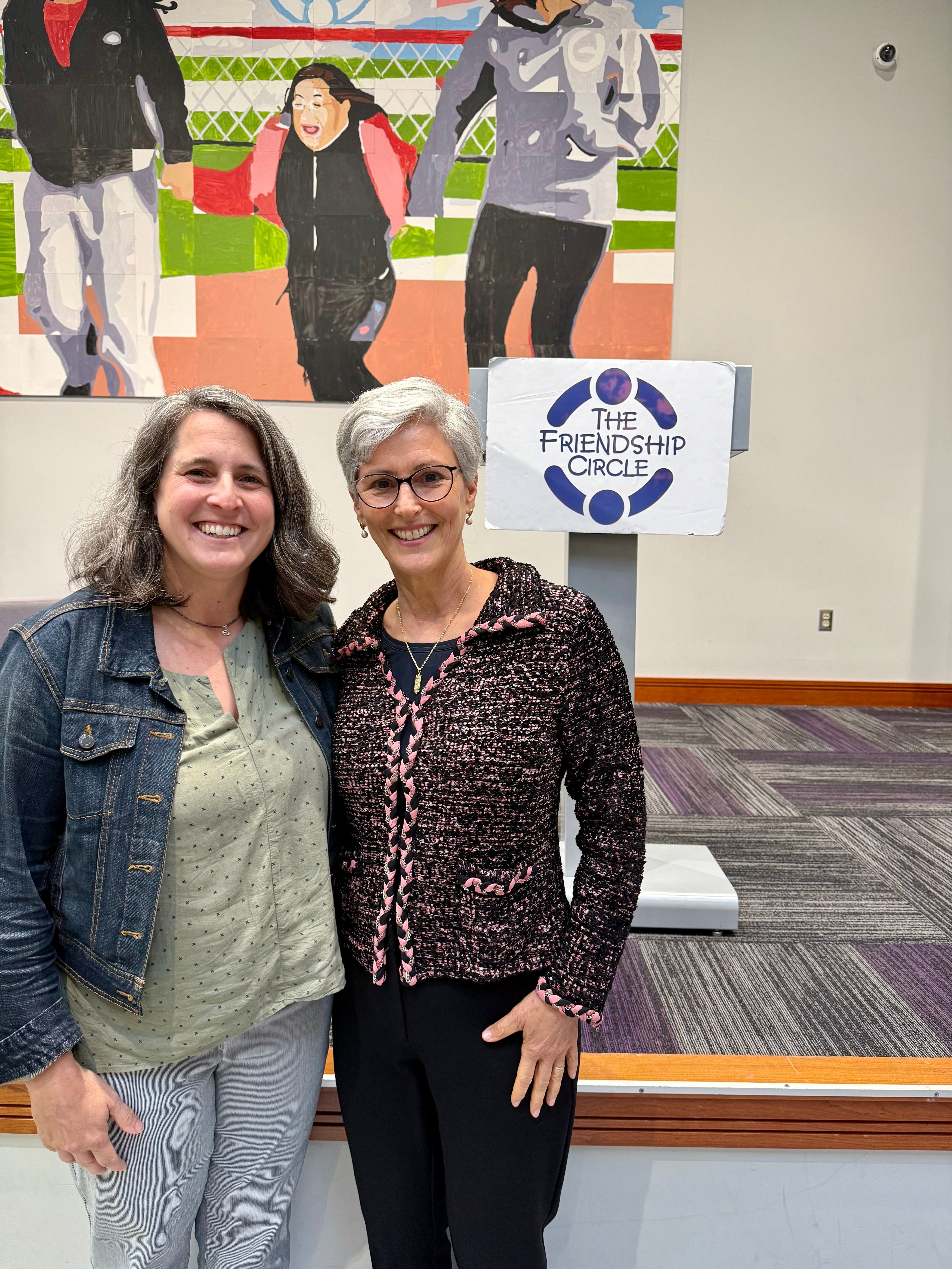 Maggie Feinstein and Rabbi Suzanne Offit stand smiling together at Friendship Circle following a community fireside chat on loss, support and hope. A Friendship Circle sign and colorful artwork are visible in the background.