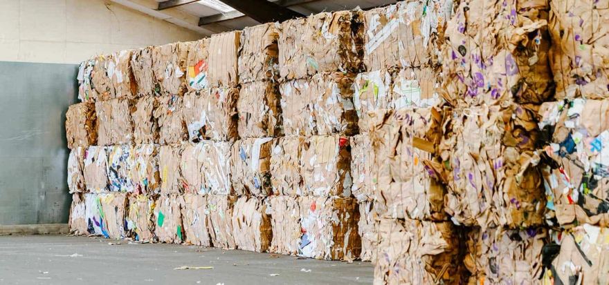 Baled cardboard stacked four high in a warehouse, the compressed bales showing mixed corrugated and printed board