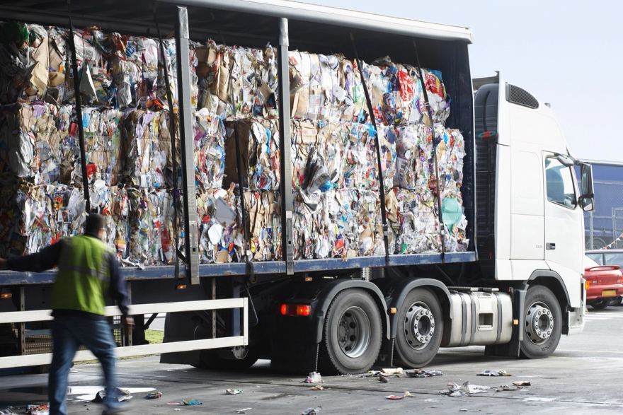 An articulated lorry with its curtain side drawn back, loaded with bales of mixed recovered paper, card and plastic stacked two high and held by webbing straps, with a worker in a hi-vis vest walking past in the foreground
