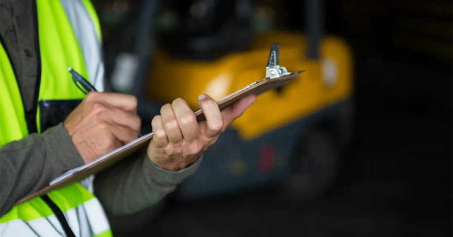 Close view of a person in a hi-vis vest writing on a metal clipboard, with a yellow forklift out of focus behind them in a warehouse