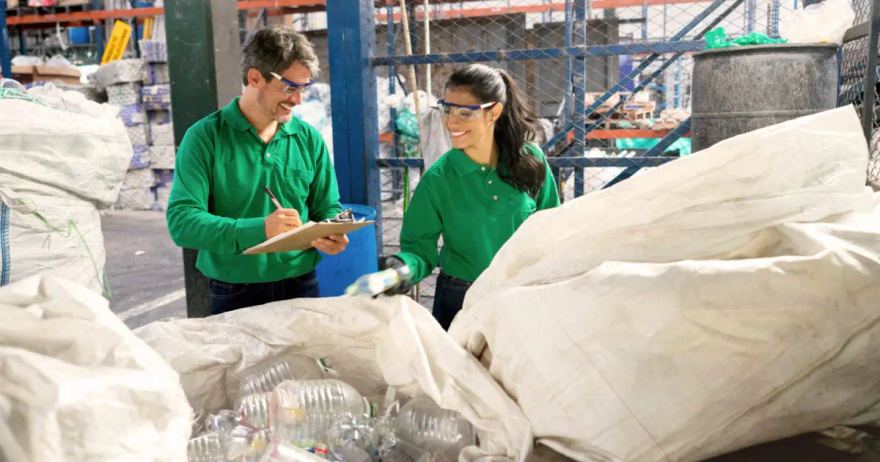 Two workers in green polo shirts and safety glasses inspecting recovered material in a sorting facility, one writing on a clipboard, with bulk bags of clear plastic bottles in the foreground