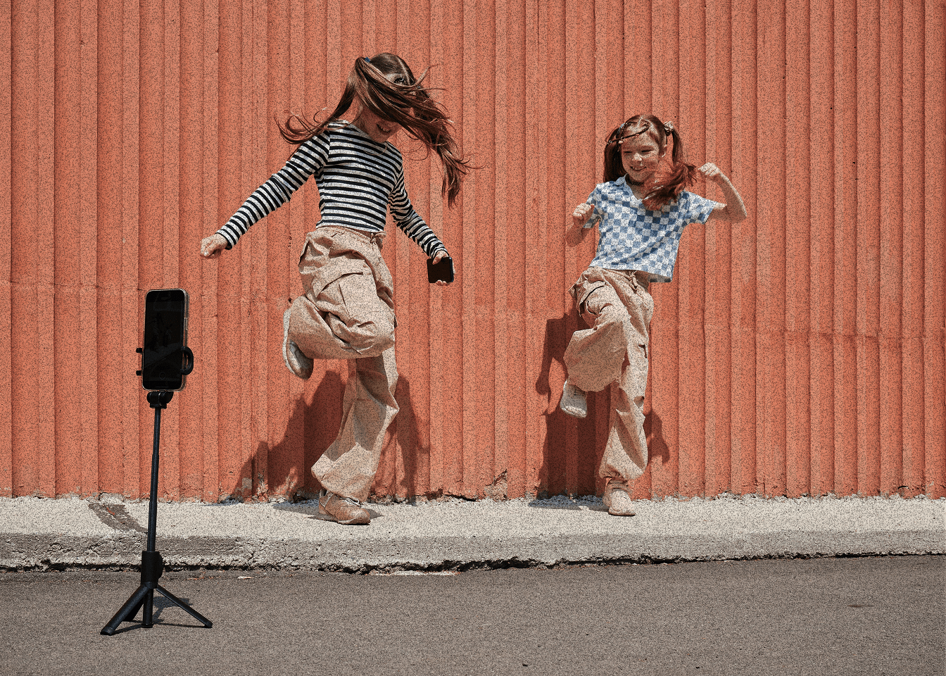 Two girls dancing in front of an orange wall while a phone on a tripod films them
