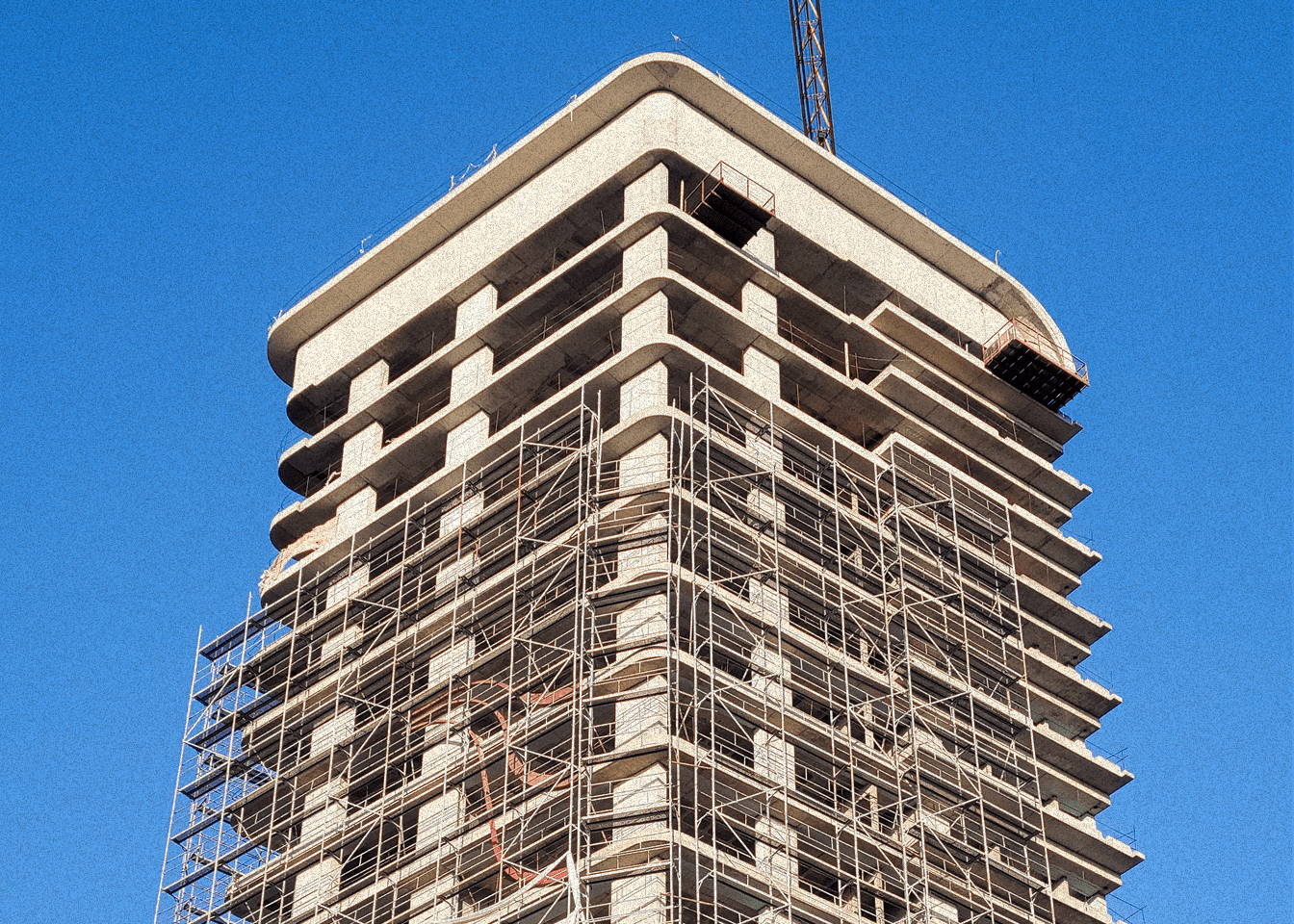 A high-rise residential tower under construction, wrapped in scaffolding against a clear blue sky