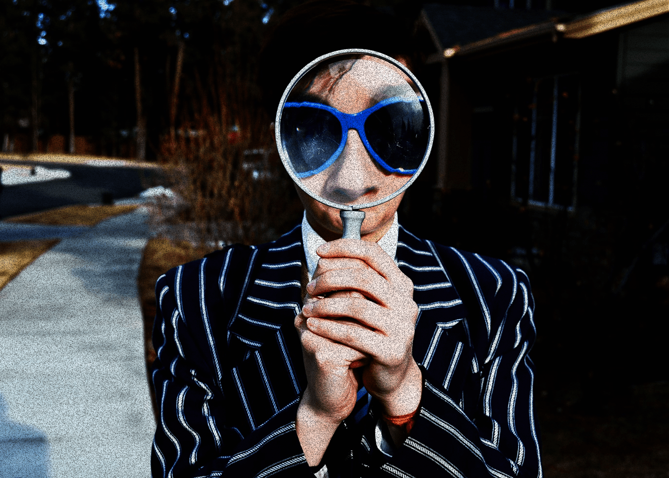 A person in a pinstriped suit and blue sunglasses holding a magnifying glass up to their face