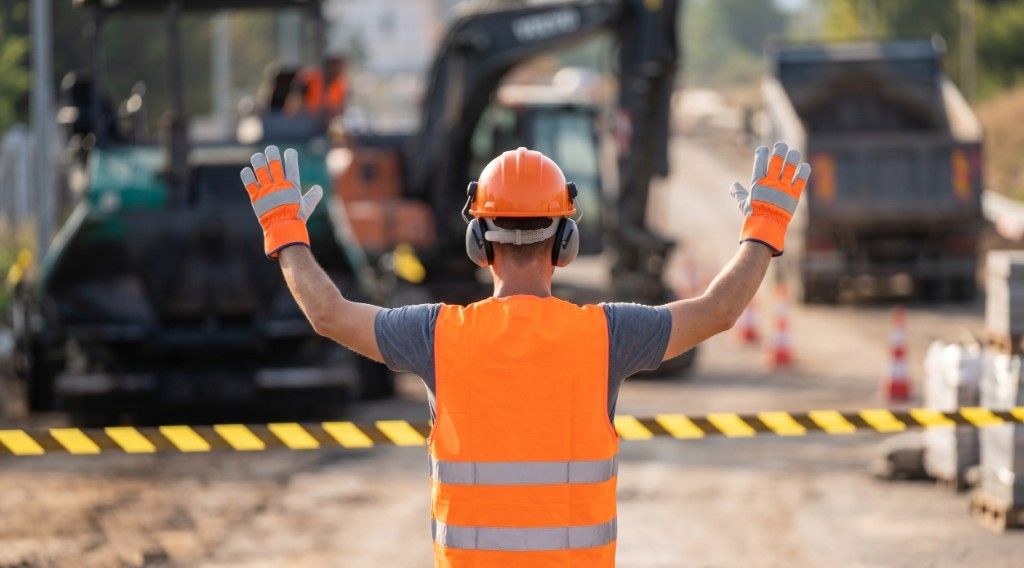 Construction worker in an orange hard hat and safety vest signaling stop behind caution tape on an active jobsite.