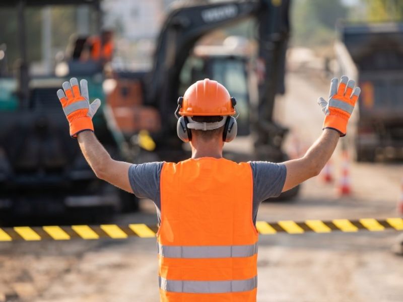 Construction worker in an orange hard hat and safety vest signaling stop behind caution tape on an active jobsite.