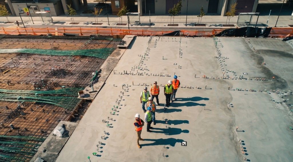 Construction team in hard hats and safety vests standing together on a foundation slab at an active jobsite.