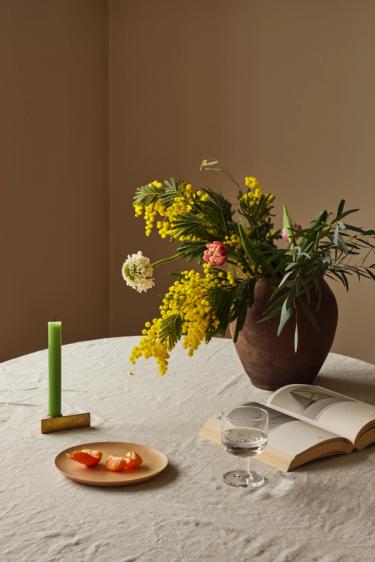 En krukke med blomster står på ett bord med duk, stearinlys, bok, glass og noen båter mandarin på en tallerken.
