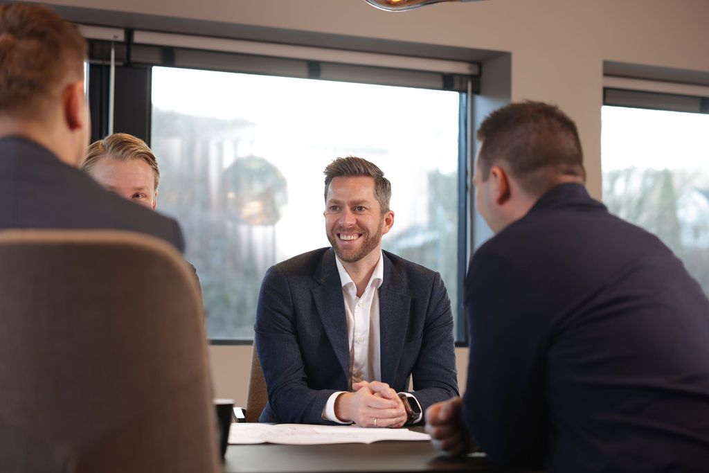 Fire menn rundt et bord og vi ser en mann smile mot kollega 