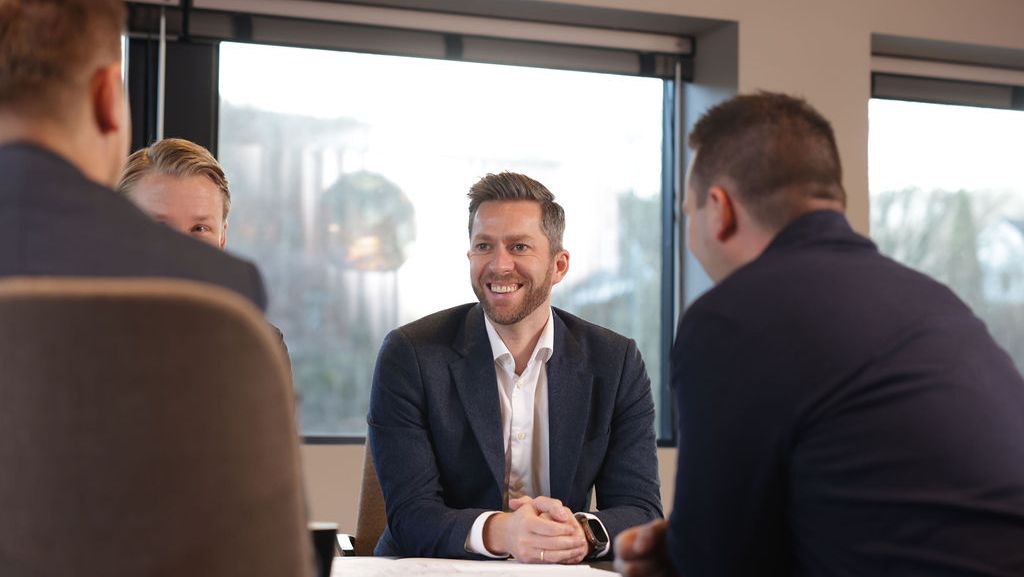 Fire menn rundt et bord og vi ser en mann smile mot kollega