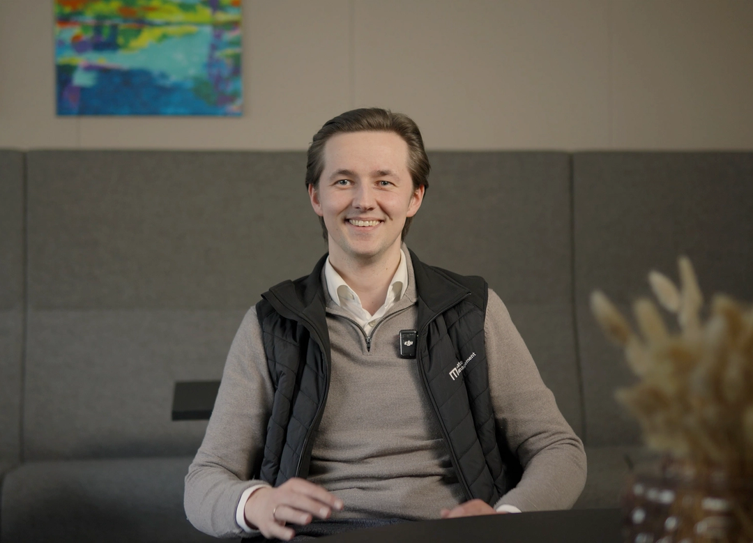 Mann smiler under videointervju, sittende på en stol med et bord foran seg