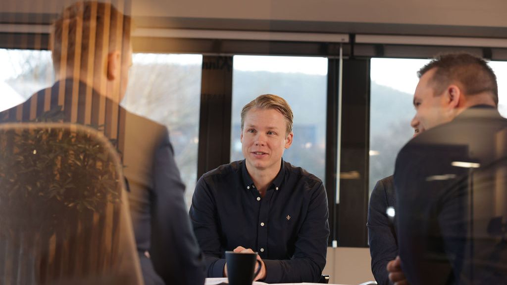 Tre menn rundt et bord hvor en person sitter mot kamera