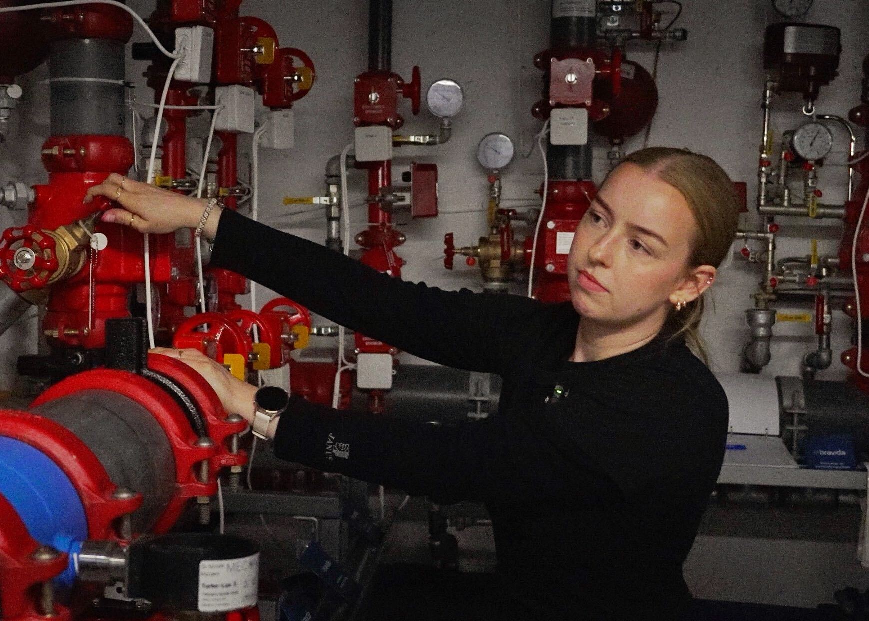 En byggdrifter i svart genser justerer en ventil på et rødt sprinkler- og rørsystem i et teknisk rom.