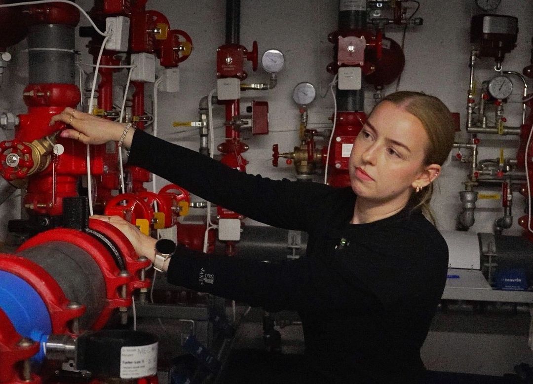 En byggdrifter i svart genser justerer en ventil på et rødt sprinkler- og rørsystem i et teknisk rom.