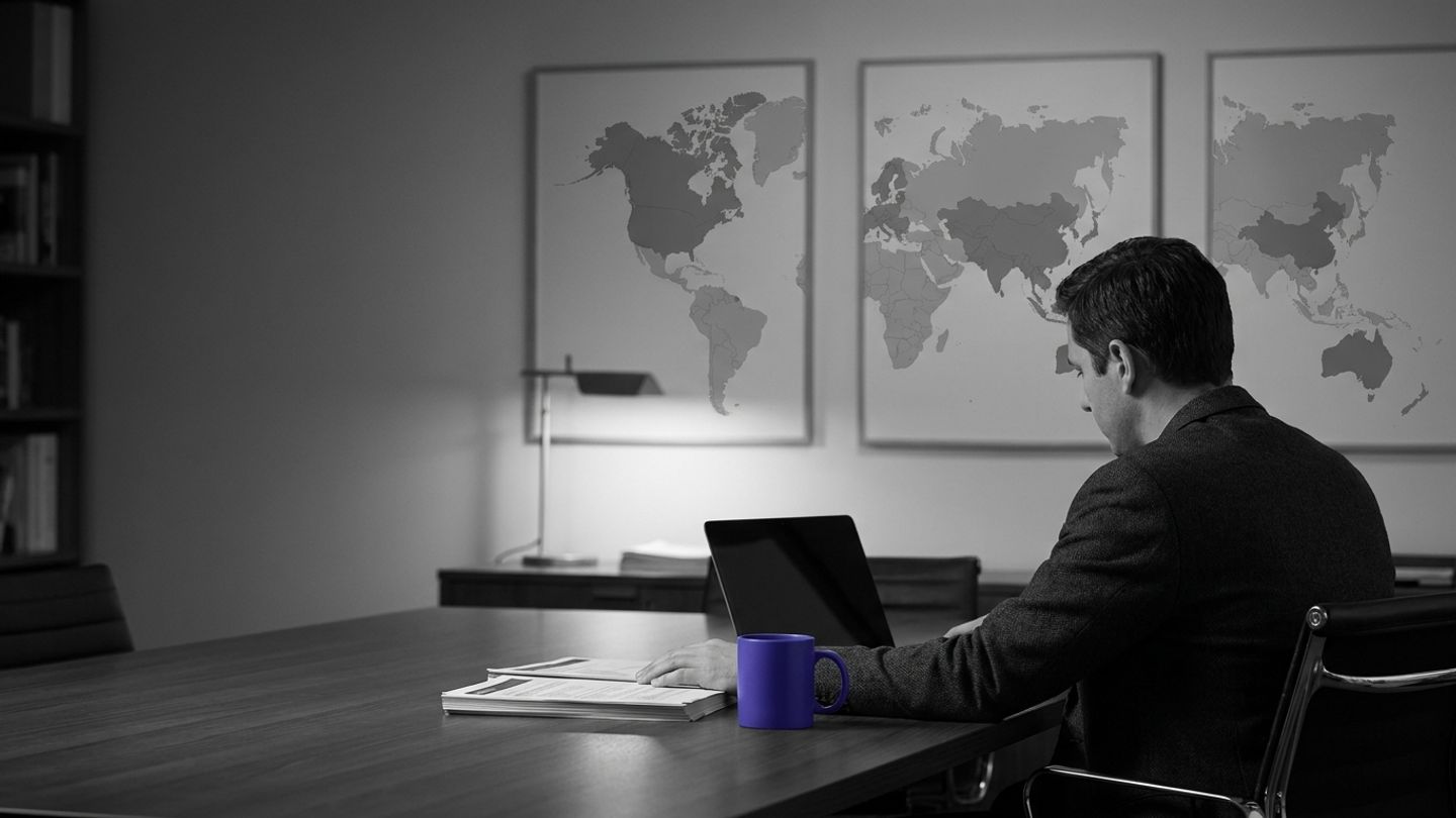 Policy analyst working at a desk beneath three abstract world maps, with a single indigo coffee mug