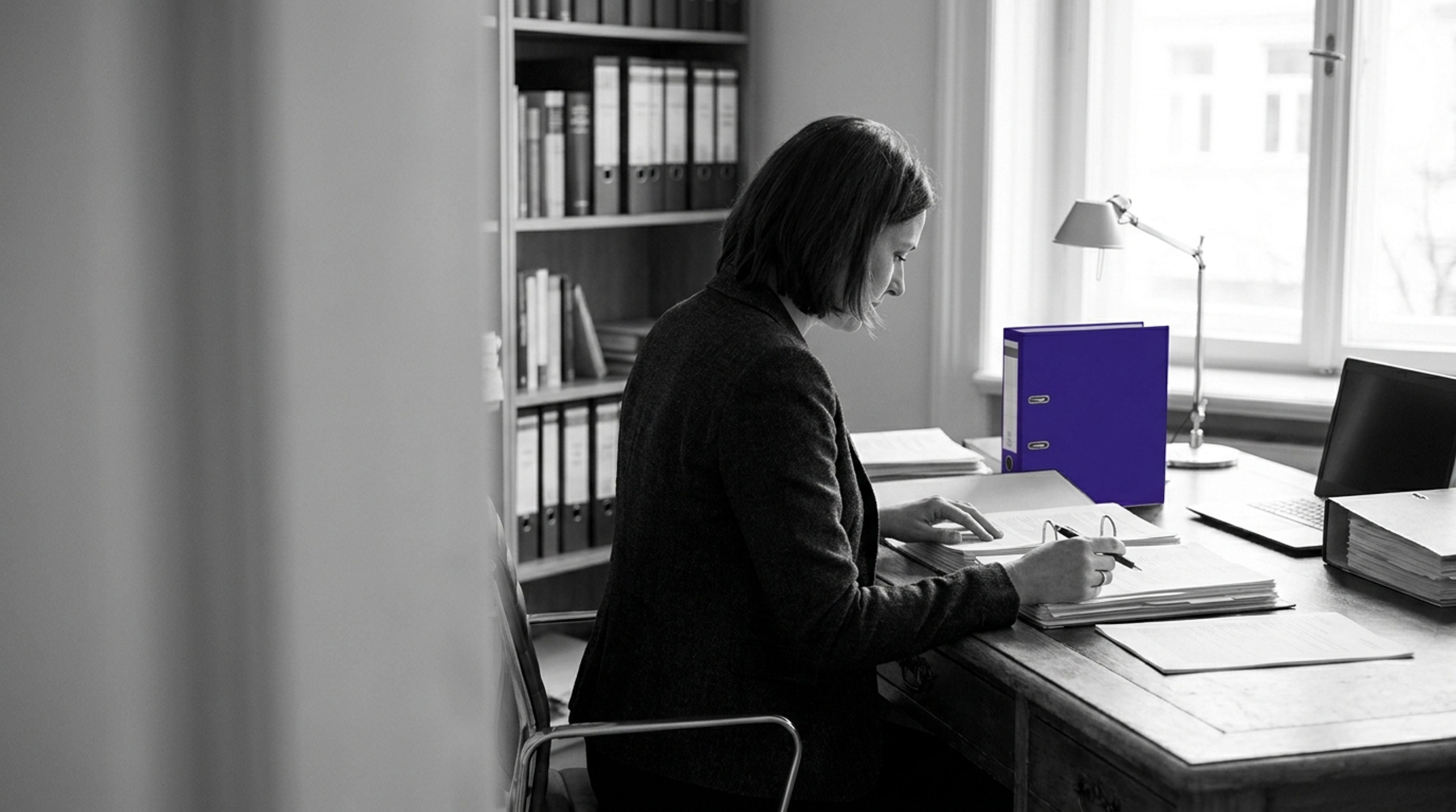 Compliance lead reviewing technical documents and binders at a desk, with a single indigo file spine